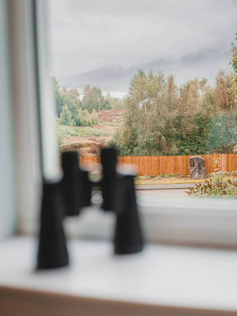 View from a window overlooking a landscape of the Scottish Highlands with trees, a wooden fence, and misty hills, binoculars resting on the inner sill.
