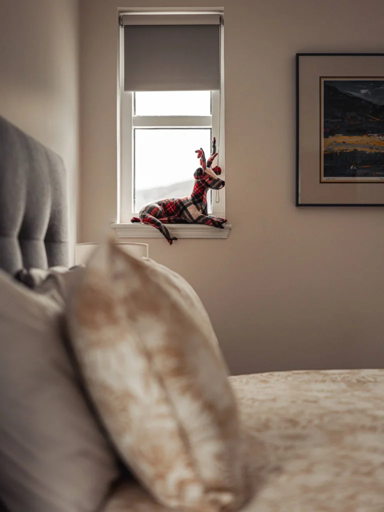 Cosy room with a bed in the foreground, a Scottish fabric stuffed deer placed on the windowsill, and a painting hanging on the wall. Warm atmosphere typical of accommodation in the Scottish Highlands.