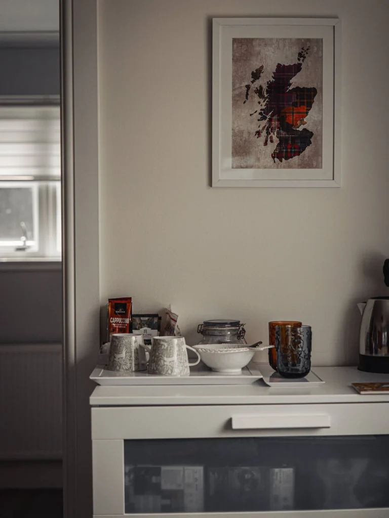Coffee corner in a rental interior in Glencoe, Highlands, with a framed map of Scotland hanging on the wall and a tray containing cups, kettle, and hot drink sachets.