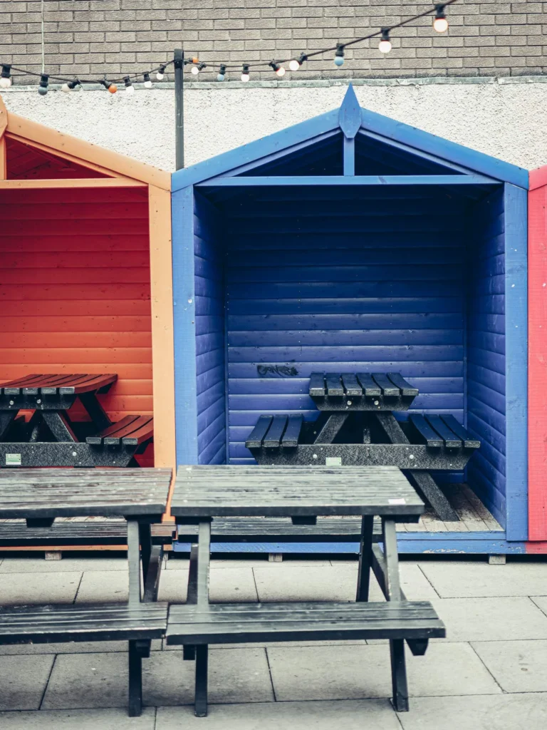 Wooden picnic tables inside the painted huts in the courtyard at Slanj-A-Va, Stirling, strung with fairy lights.