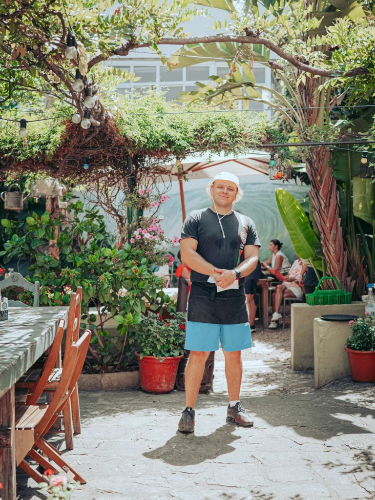 The garden of the restaurant The Garden Bar and Grill in Lagos, its numerous wooden tables and its green pergola, with a staff member standing in the middle.