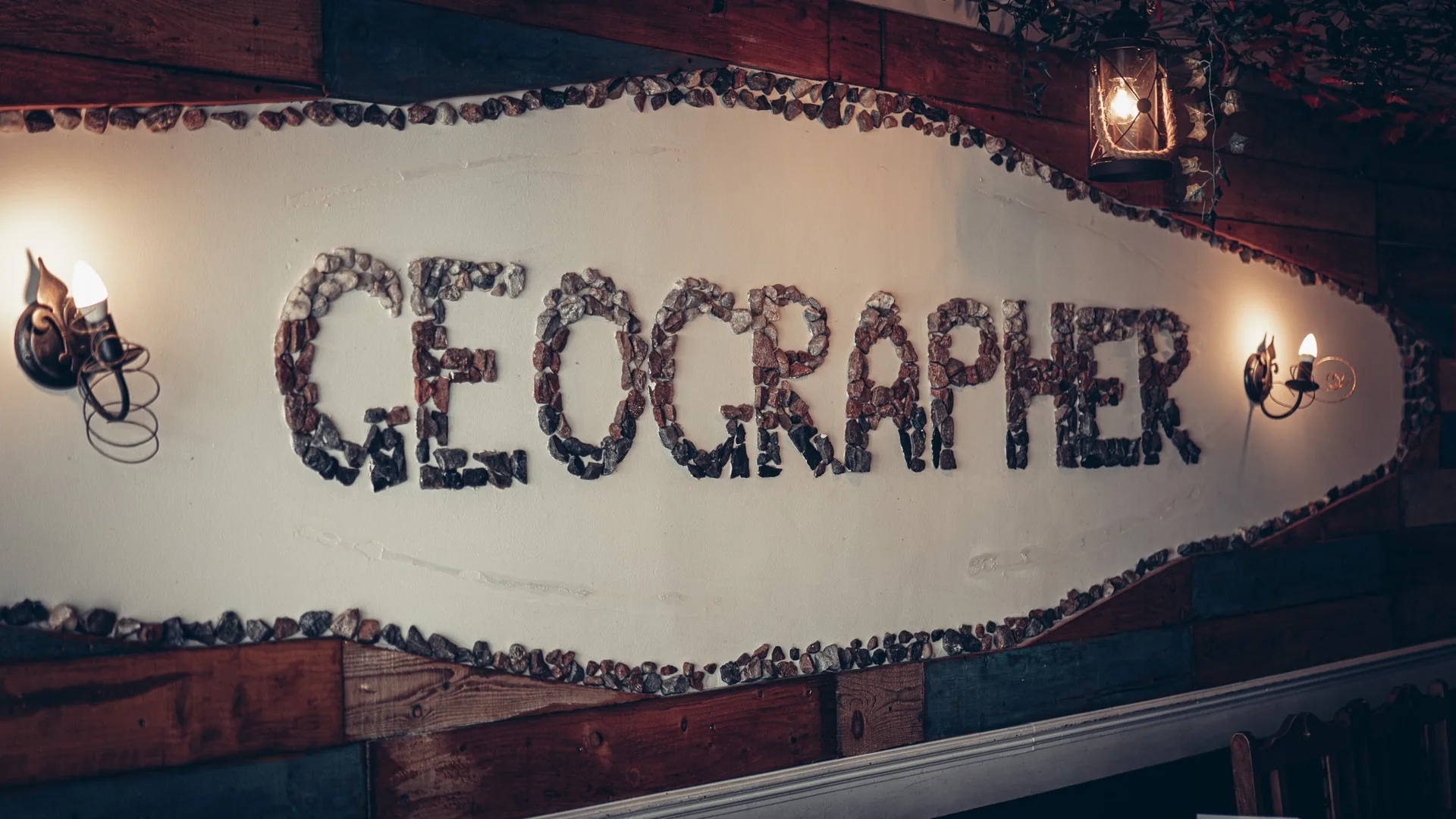 The name of The Geographer spelled out in pebbles on a panelled wall in Fort William, lit by warm wall lights.