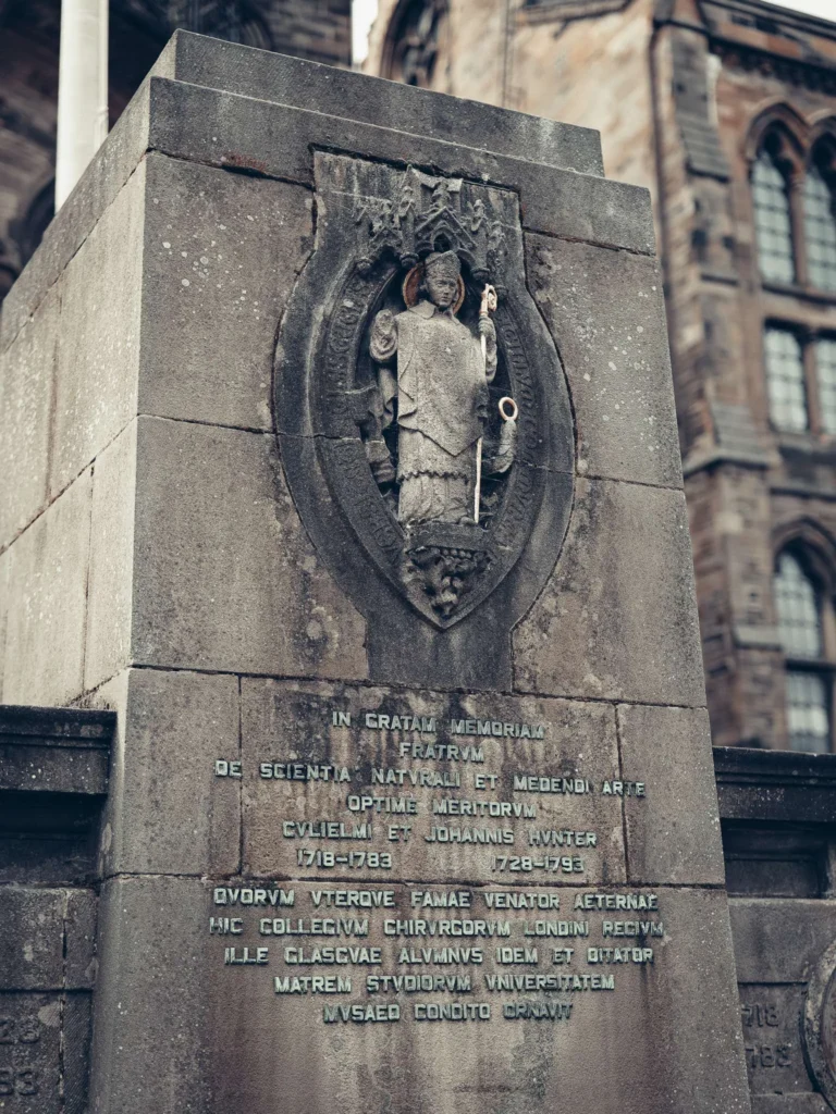 Stone memorial monument, relief statue and Latin inscription, in front of a neo-Gothic facade of the University of Glasgow.