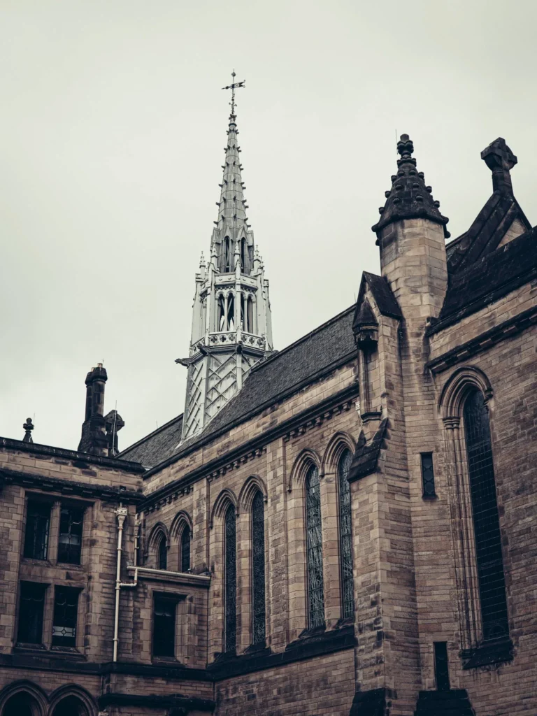 Stone facade of the University of Glasgow topped with a large spire bearing a cross: the Gothic Revival architecture by George Gilbert Scott is as much cathedral as it is university.