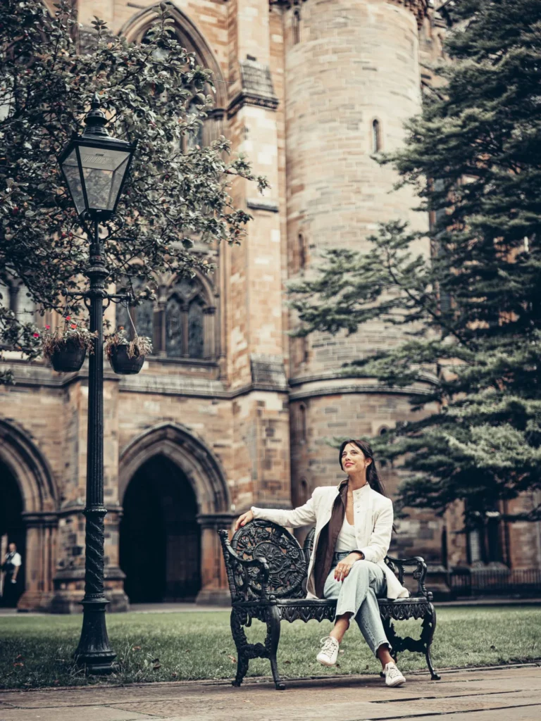 Caroline sitting on a wrought iron bench in an inner courtyard of the University of Glasgow, in front of the stone facade and its arches.