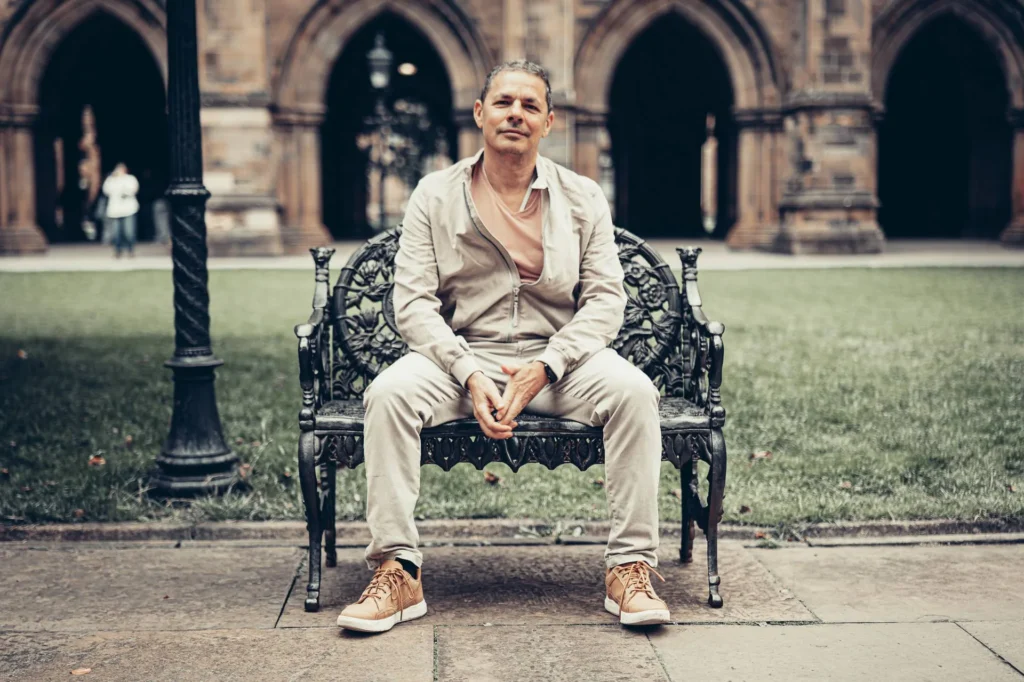 Xavier sitting on a wrought iron bench in a quadrangle at the University of Glasgow, the arches behind him.