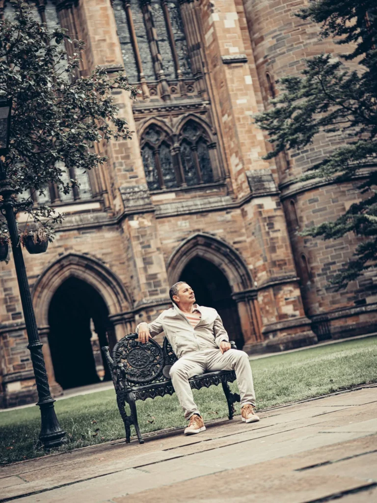 Xavier sitting on a bench in front of an archway and stained glass facade of the University of Glasgow, in a green courtyard.