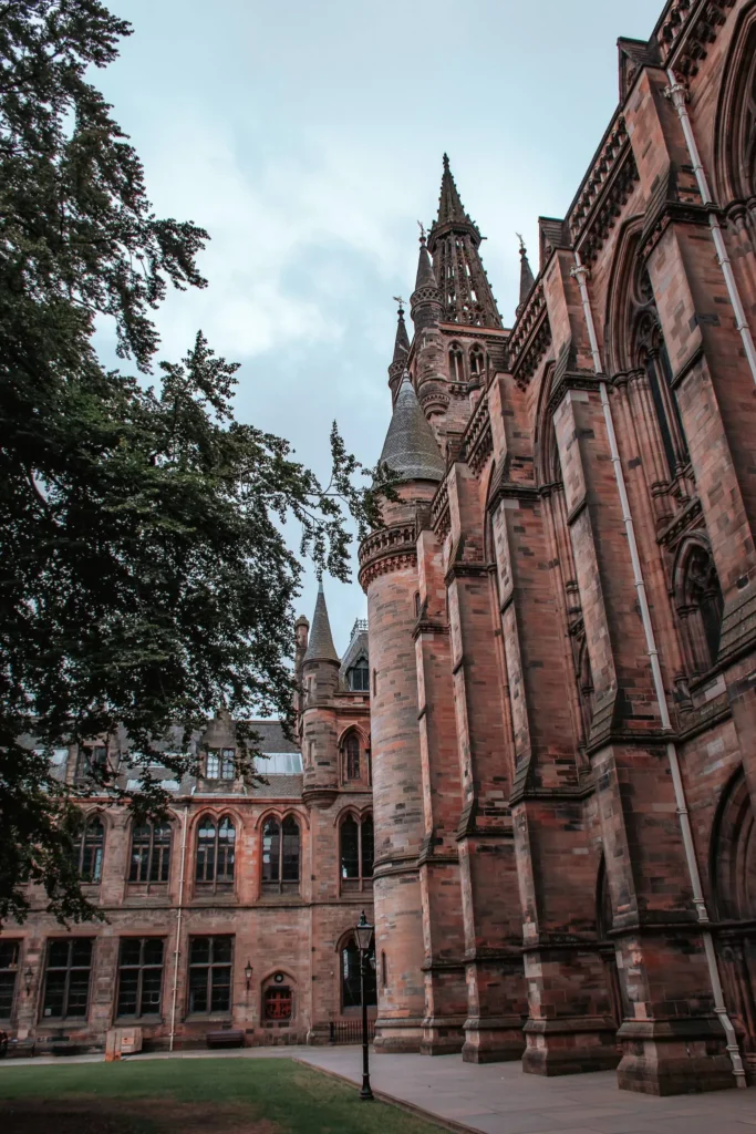 Gothic facade of the University of Glasgow in Scotland, with its characteristic towers and arches, photographed on a cloudy summer day.