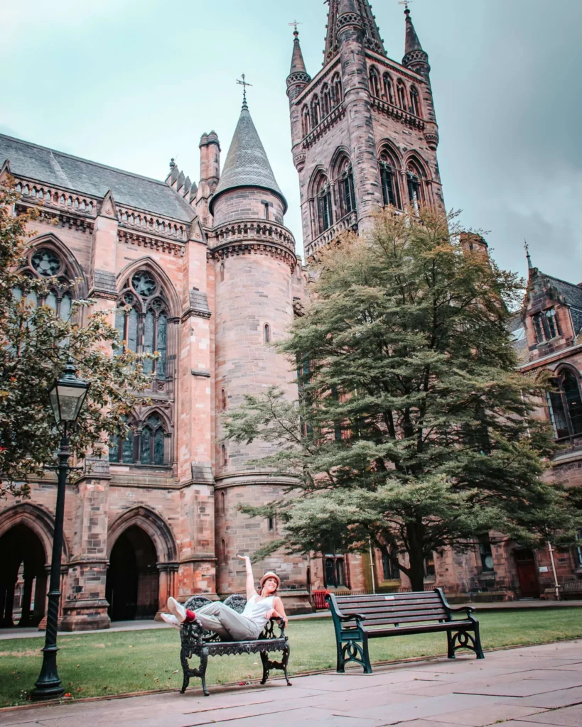 Caroline is lying on a bench in front of the Gothic architecture of the University of Glasgow, Scotland, on a cloudy day.