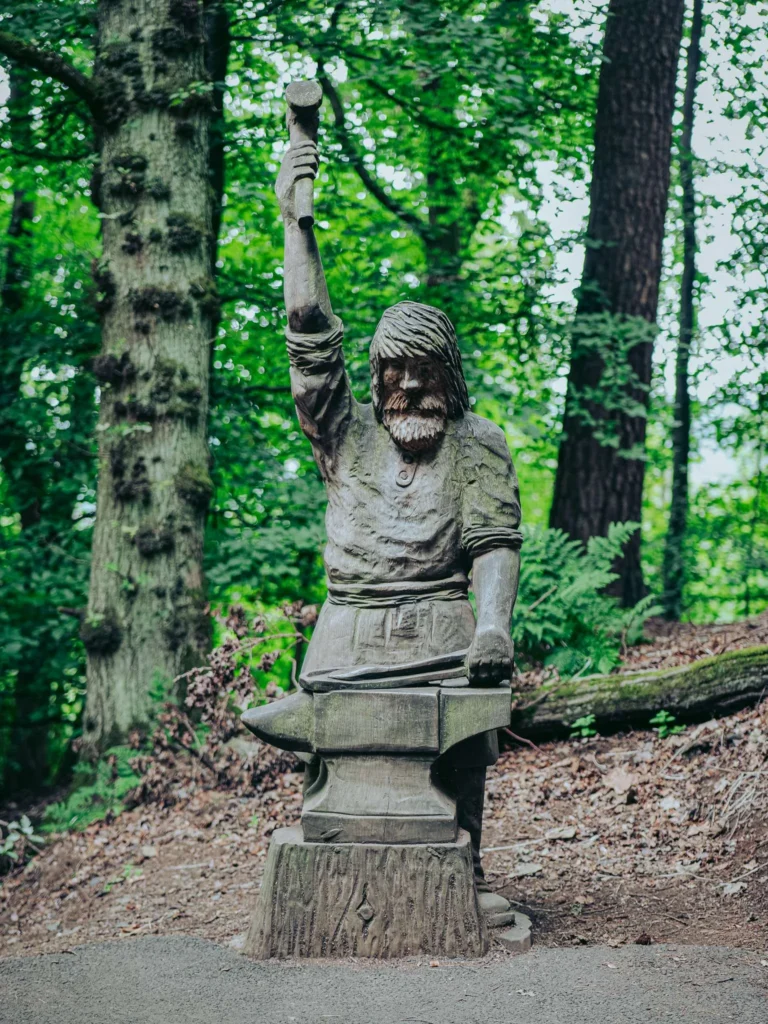 Wooden statue depicting a blacksmith with a raised hammer, located in a forested environment near the Wallace Monument in Stirling, Scotland in August 2026.