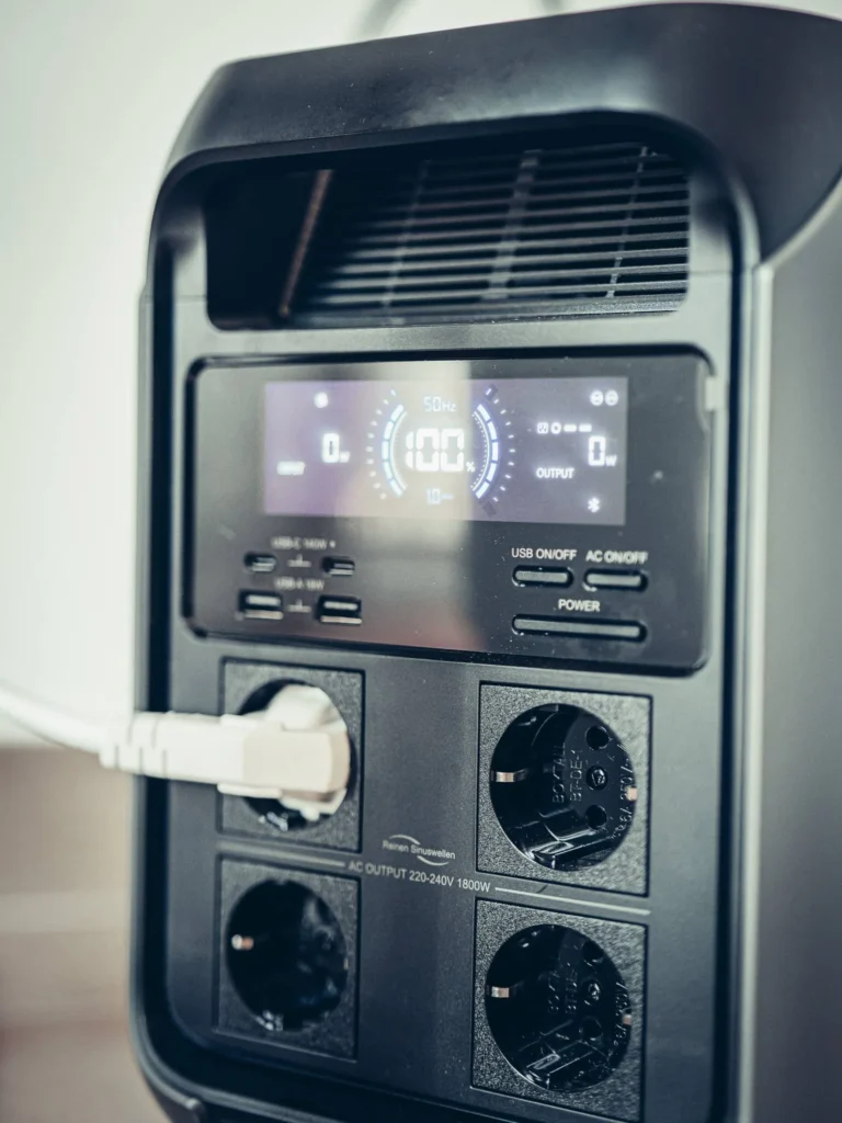 Close-up of the ALLPOWERS P1800 power station being tested, its integrated color screen showing 100% battery, with the four AC outlets, USB ports, and 12V car cigarette lighter socket visible on the front.