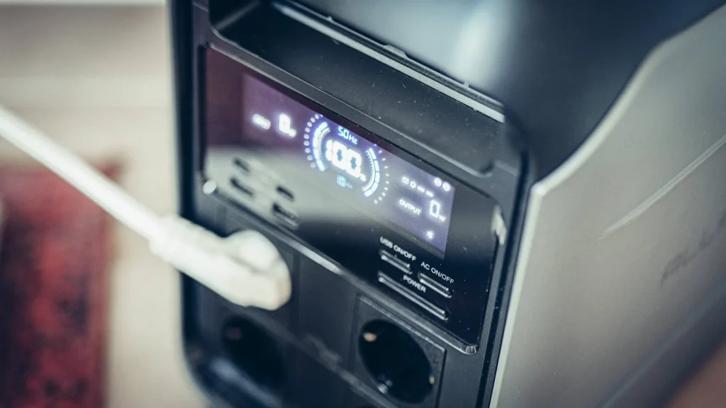 Close-up of the ALLPOWERS P1800 power station under test, showing its integrated color screen, several visible ports (USB, USB-C, power outlets), and a plugged-in socket. The screen displays the charge status and output information.