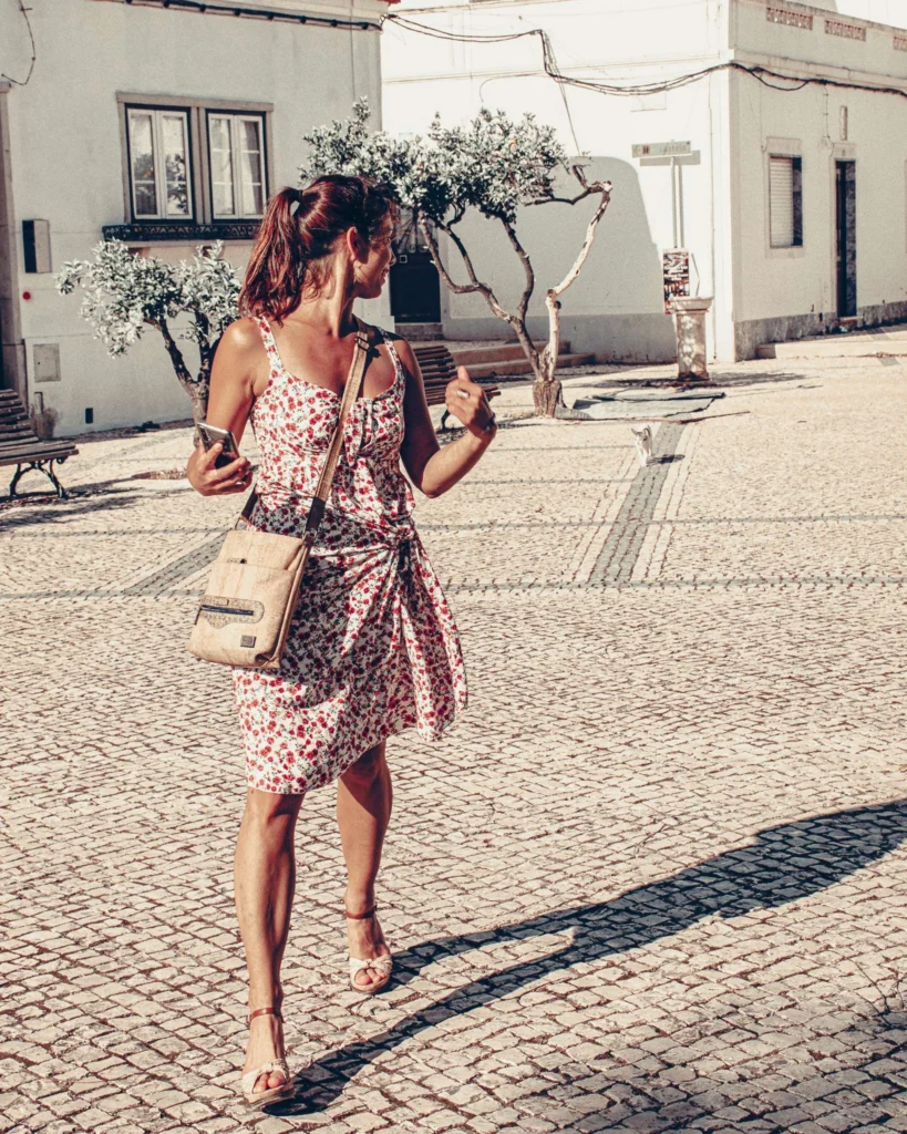 Caroline walks on a sunny cobblestone square in Castro Marim, surrounded by white buildings and olive trees, in the Algarve region of Portugal.