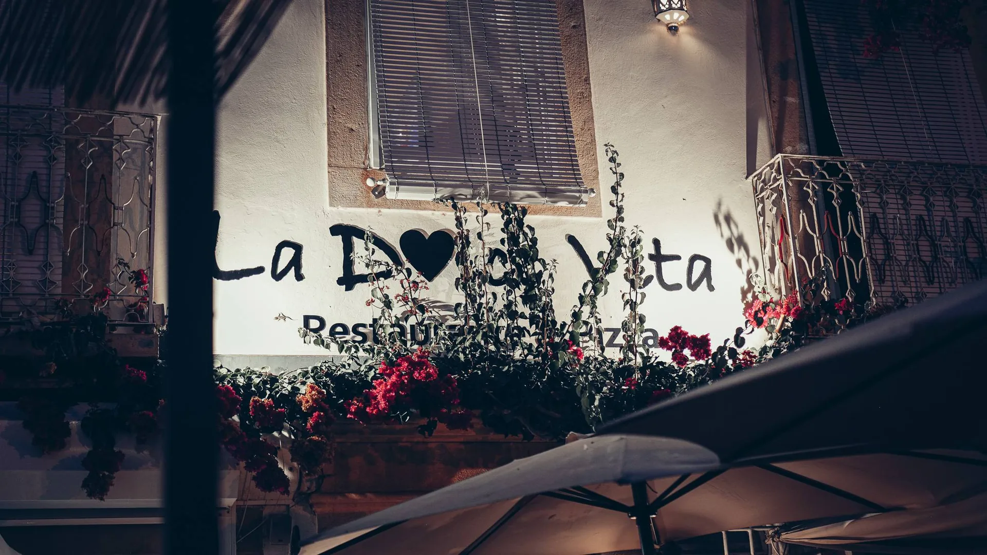 Night facade of the pizzeria restaurant Dolce Vita in Tavira, Algarve, with illuminated sign and flowered balcony.
