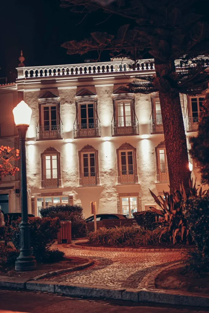 Facade of an elegant building illuminated at night, with balustrade windows and a streetlamp, in a cobblestone street surrounded by greenery.