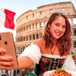 Caroline taking a selfie in front of the Colosseum in Rome with a plate of pasta and an Italian flag in the background.