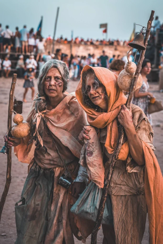 Two people dressed as medieval characters stroll during the medieval festival of Castro Marim, with a crowd and ramparts in the background.