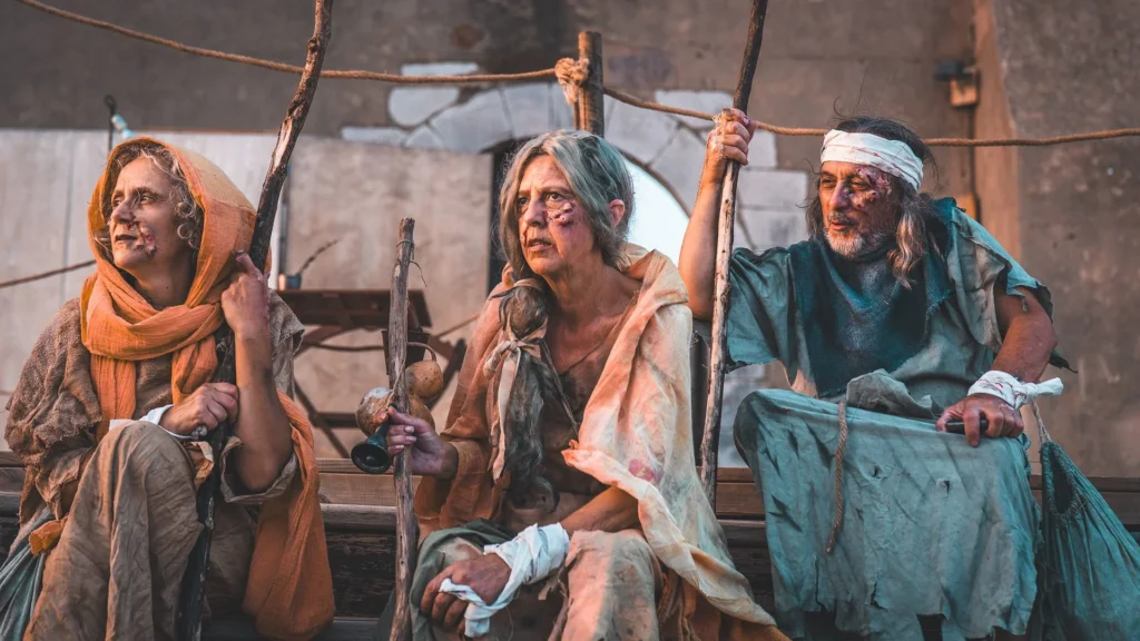 Three people dressed as medieval characters sitting on a platform during a historical reenactment, wearing worn costumes and rustic accessories.