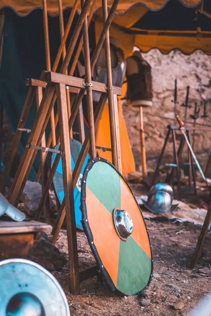 Colored medieval shields, helmets, and spears displayed under a tent during an outdoor historical reenactment.