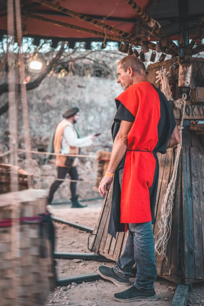 Adult man in a red and black medieval costume standing near a wooden structure during an outdoor show, another costumed man in the background.