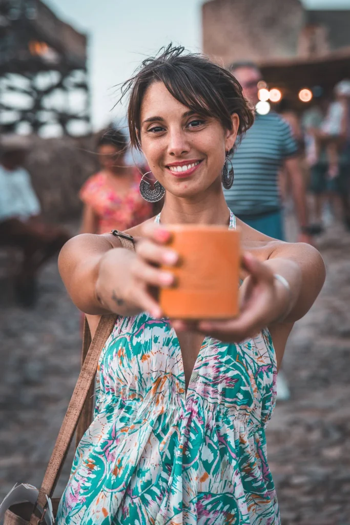 Caroline in a floral dress smiles while holding out a cup at the medieval festival of Castro Marim, with participants and decorations in the background.