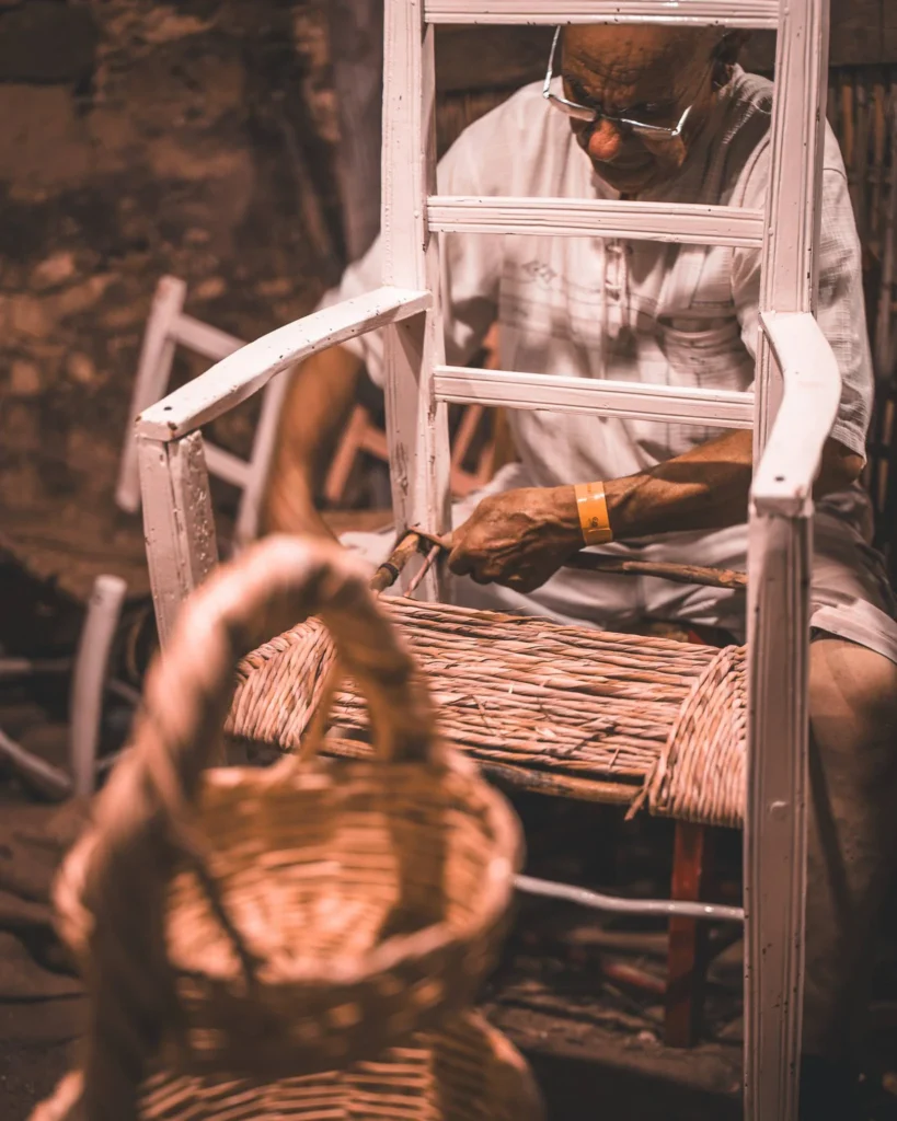 A man weaves the seat of a wicker chair during a craft demonstration, with baskets in the foreground. Medieval and artisanal atmosphere.
