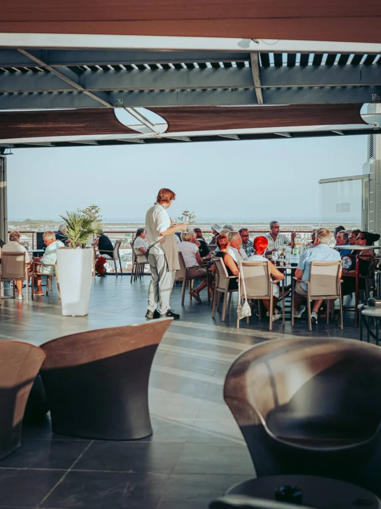 Nomad's terrace in Tavira at aperitif time: occupied tables, waiter in the room, and an unobstructed view of the Ria Formosa in the background.