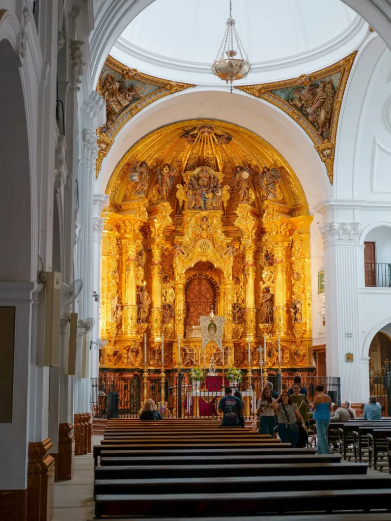 Gilded altarpiece and white interior of El Rocío sanctuary, with visitors beside the pews